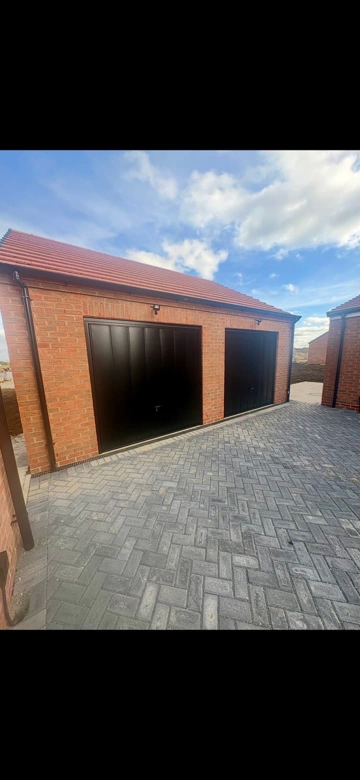 Red brick double garage with black doors and a grey herringbone paved driveway.