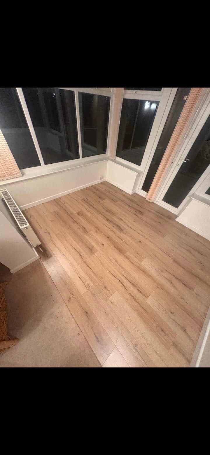 Empty sunroom with light oak laminate flooring, white walls, and large windows at night.