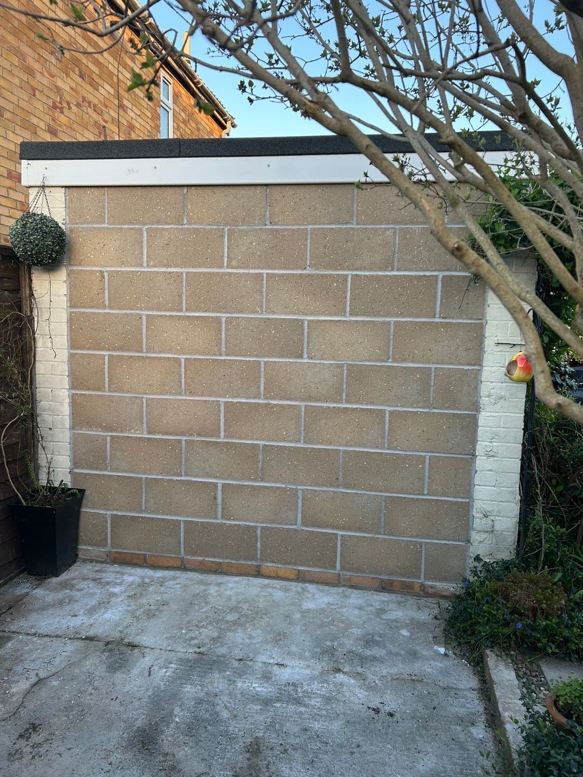 Tan concrete block wall on a concrete patio, flanked by white pillars and garden greenery.