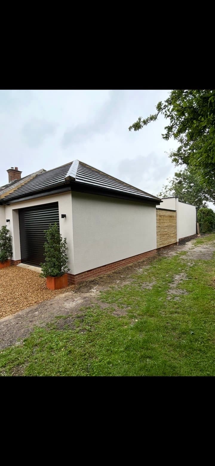 Modern white rendered garage with black roller door, dark tiled roof, and wooden fence extension.